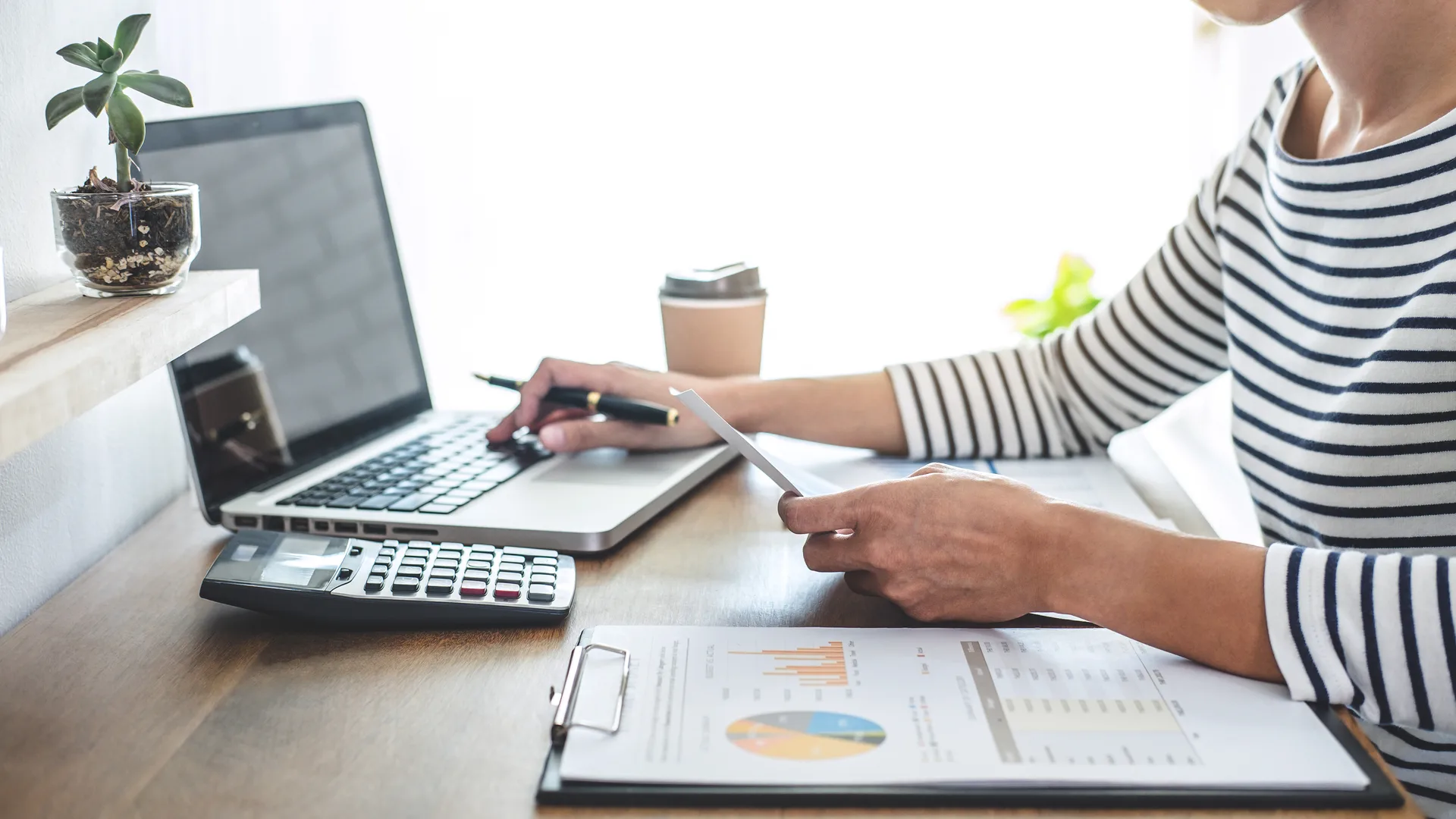 Frau am Schreibtisch arbeitet mit Laptop, Taschenrechner und Unterlagen an ihrer Finanzplanung
