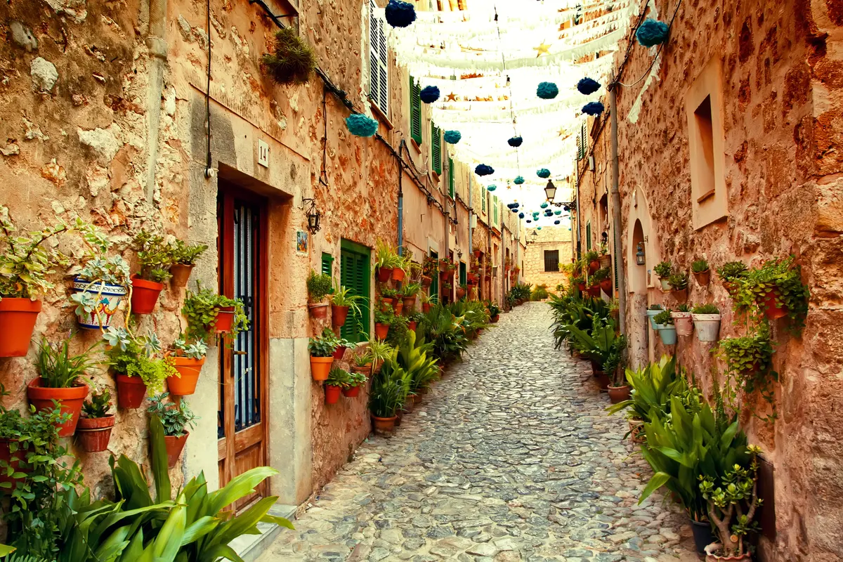 Enge Gasse in einem mallorquinischen Dorf mit Natursteinhäusern, bepflanzten Blumentöpfen und Lichterketten über dem Kopfsteinpflaster